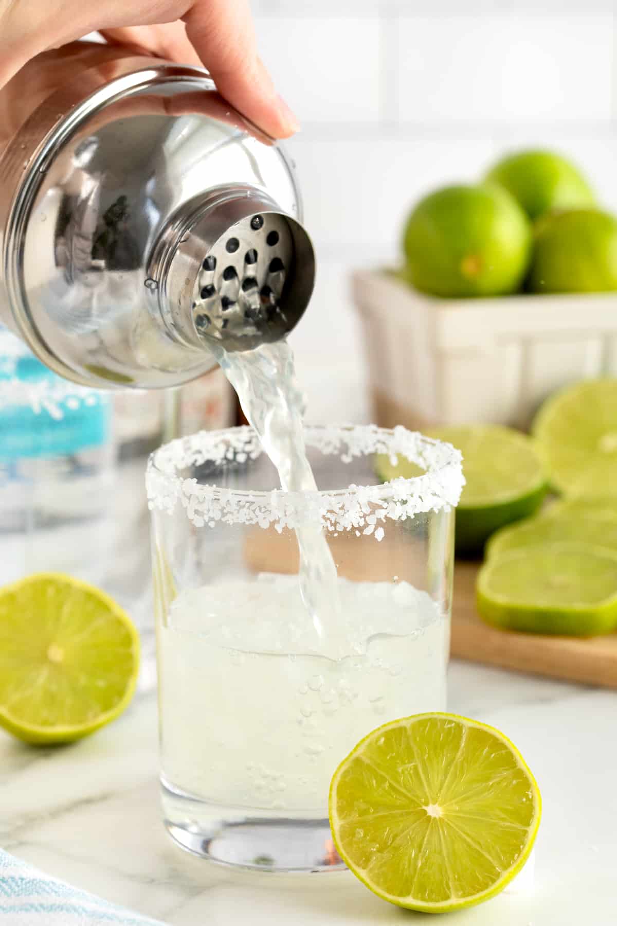 A salt rimmed glass being filled with margarita on a rimmed wooden serving tray. There is a lime wedge on the rim of the glass.