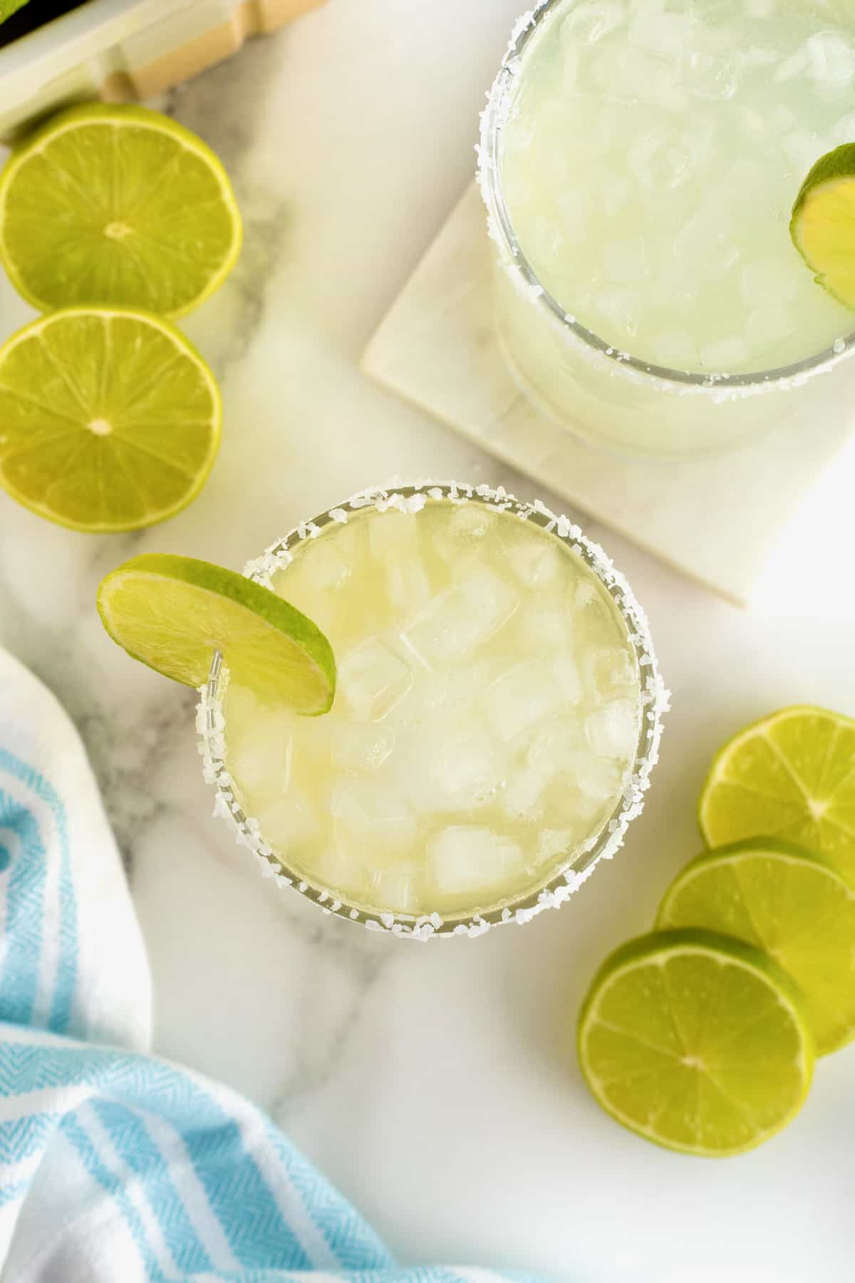 A salt rimmed glass filled with margarita. There is a lime wedge on the rim of the glass and lime slices scattered around.