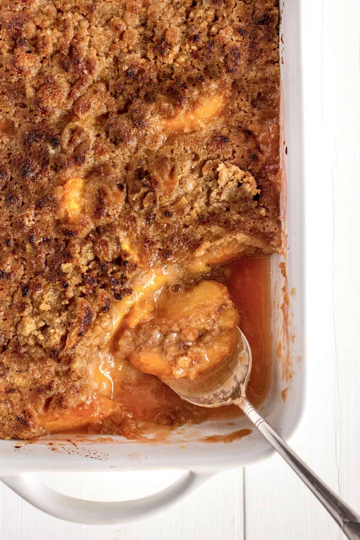 A baking dish of peach crumble with a serving removed and spoon laying in it.