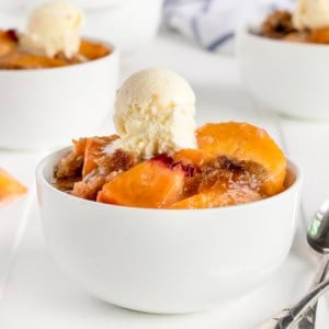 White bowl filled with peach crumble with a small scoop of vanilla ice cream on top.