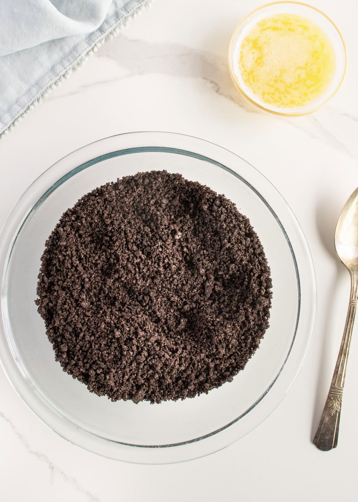 Chocolate cookie crumbs in a glass bowl next to a small white dish of melted butter.