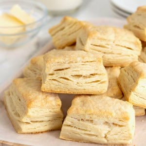 A pile of buttermilk biscuits on a white surface. Behind the biscuits is a small dish of butter.