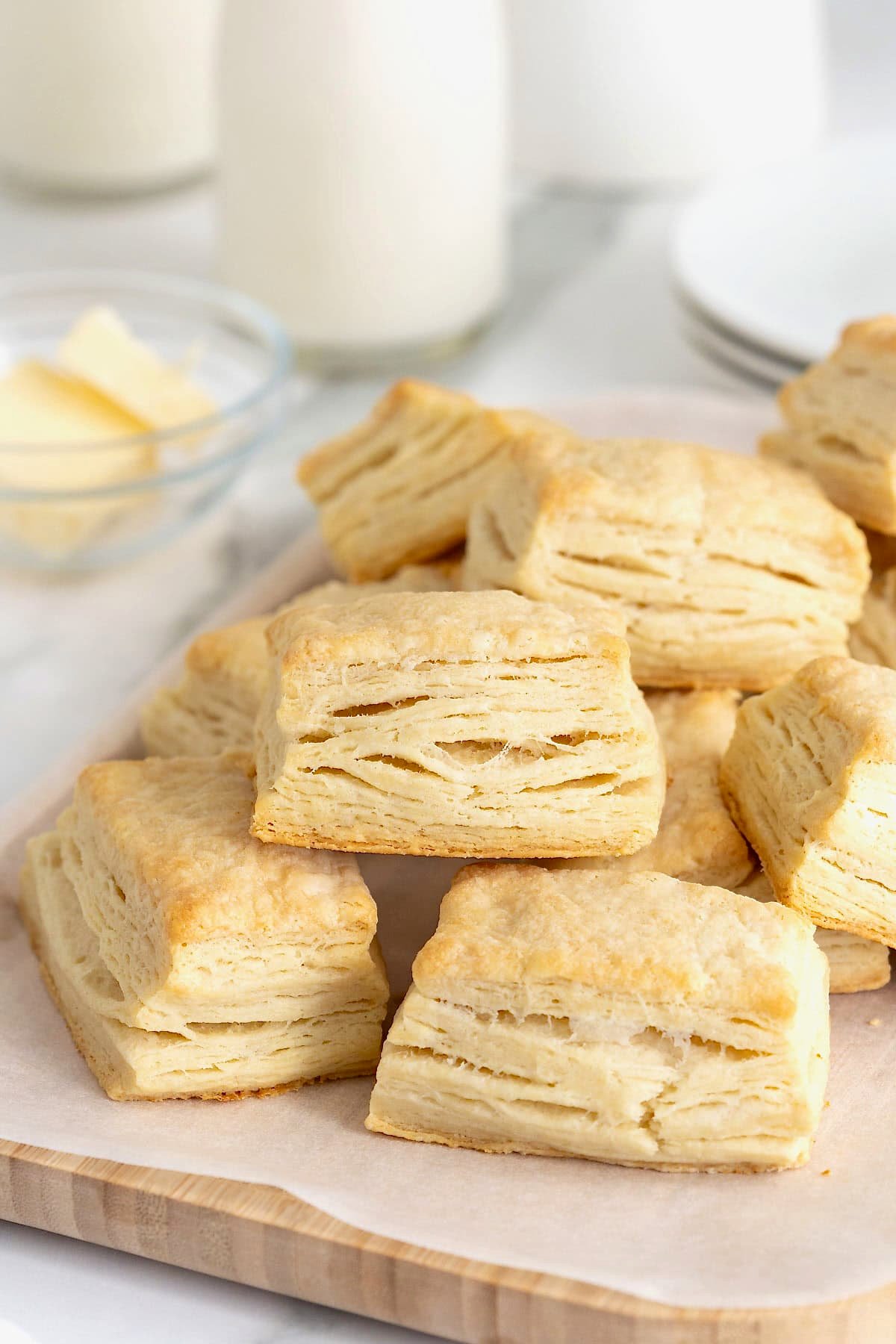 A pile of buttermilk biscuits on a white surface. Behind the biscuits is a small dish f butter.