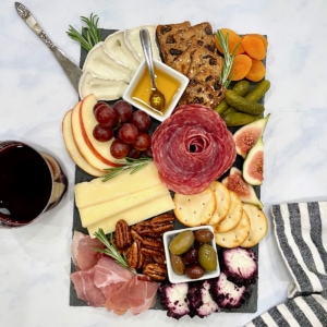 A small wooden board with meats, cheeses, apple slices, red grapes, an assortment of olives and crackers.