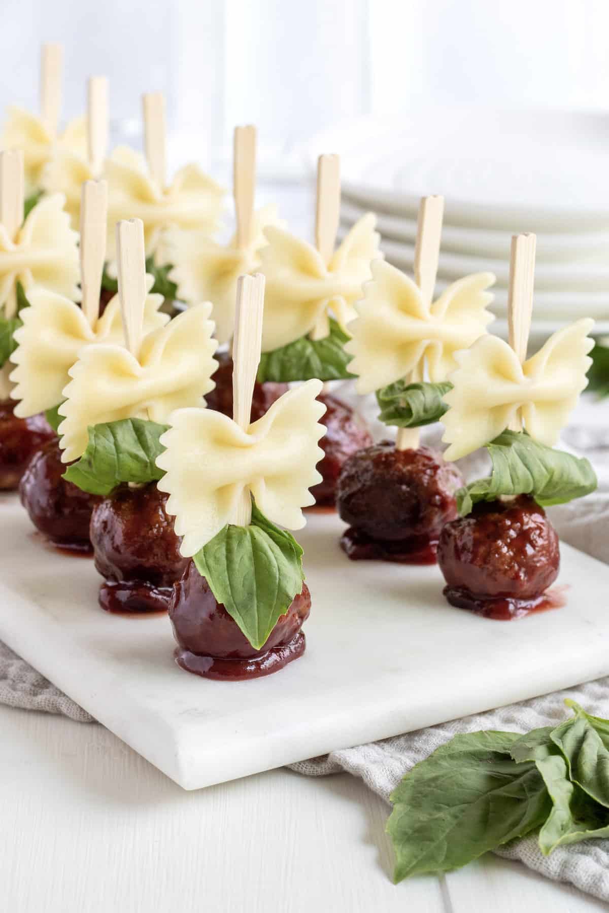 12 wooden skewers of bowtie pasta and meatballs on a white marble serving plate.