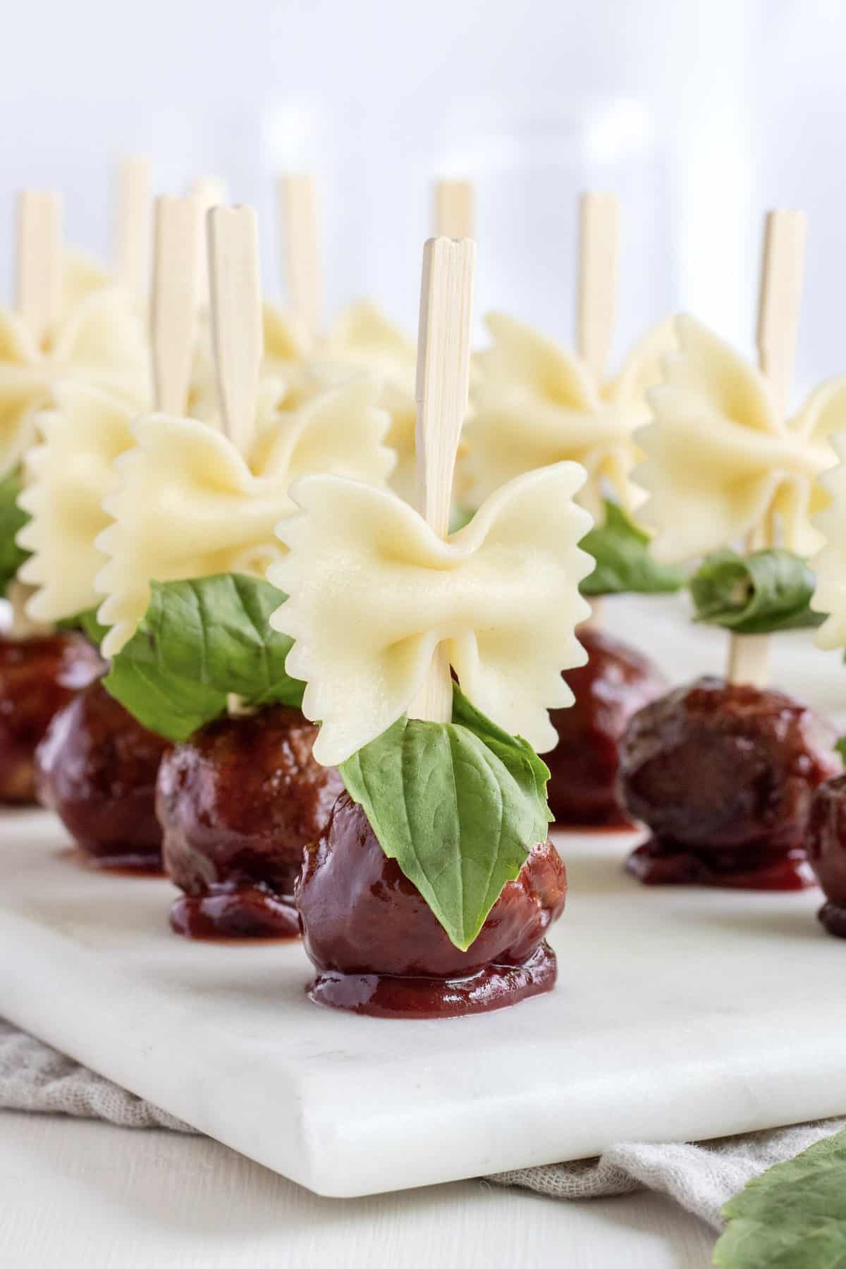 12 wooden skewers with bowtie pasta and saucy meatballs on a white marble serving tray.