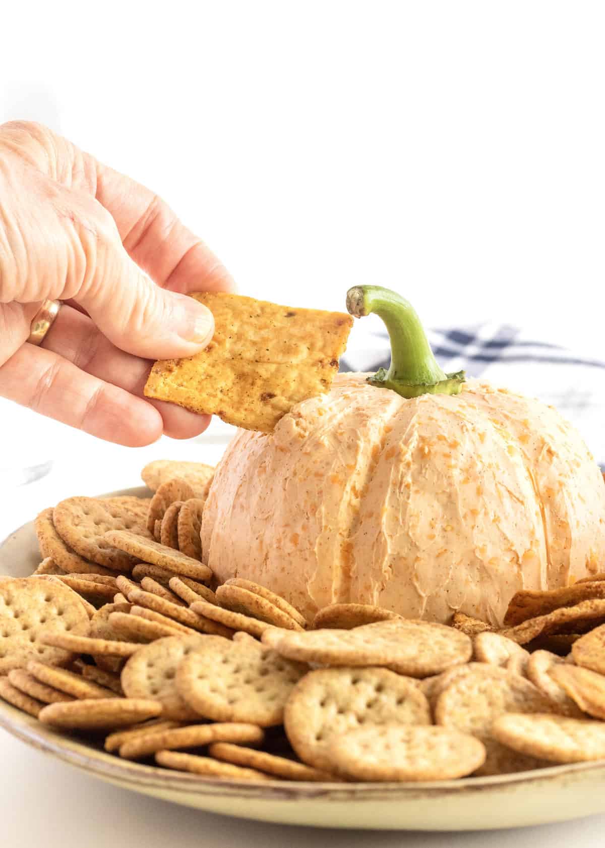 A cheese ball in the shape of a pumpkin surrounded by crackers on a large white serving plate. A hand is dipping a cracker into the cheeseball.