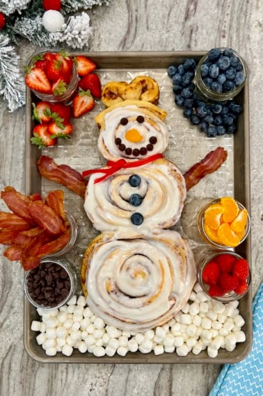 A snowman made of three cinnamon rolls on a baking sheet surrounded by mason jars filled with bacon, strawberries and mandarin slices.