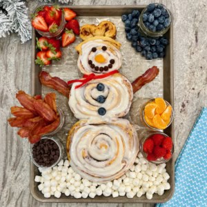 A snowman made of three cinnamon rolls on a baking sheet surrounded by mason jars filled with bacon, strawberries and mandarin slices.