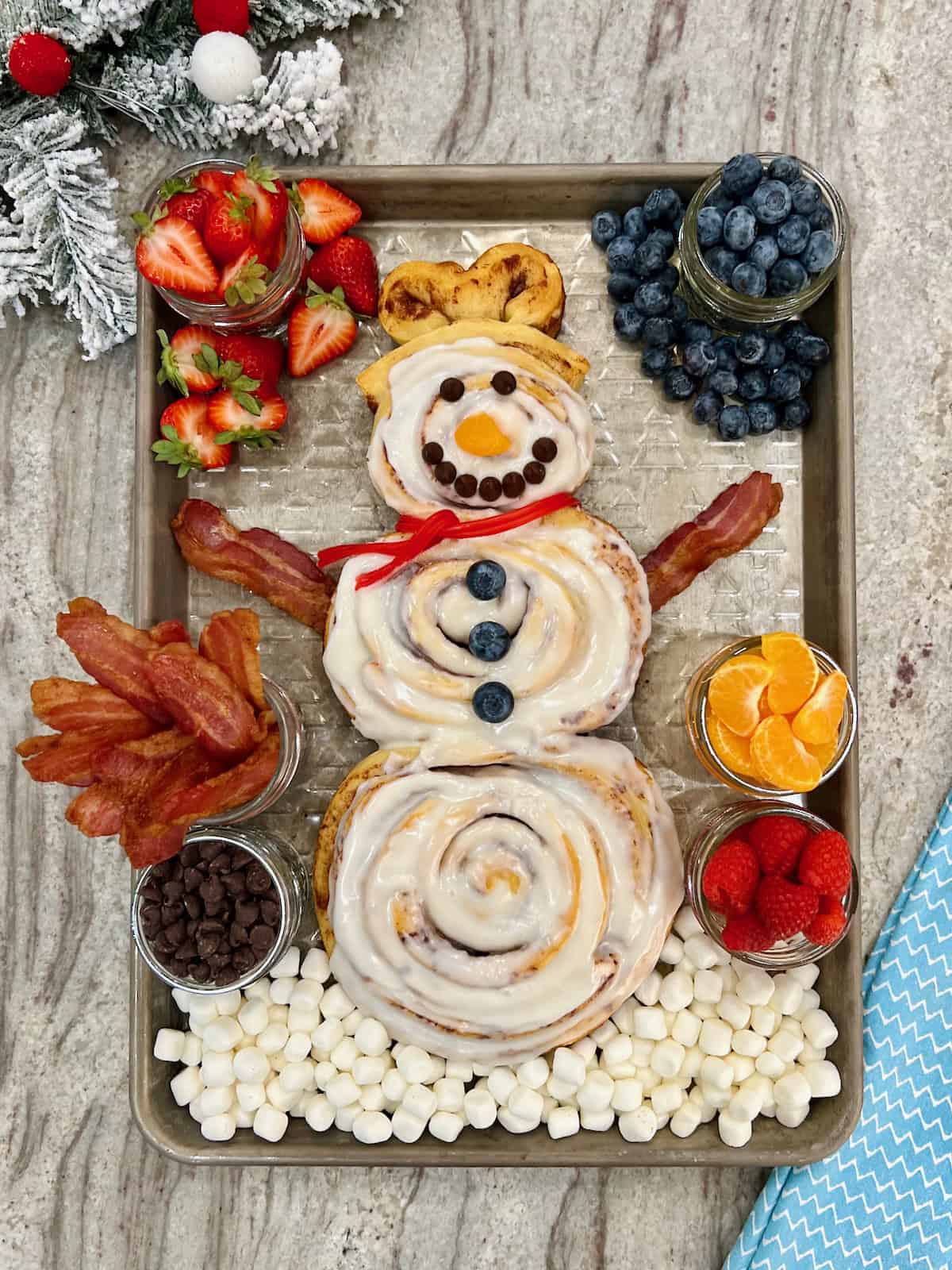A snowman made of three cinnamon rolls on a baking sheet surrounded by mason jars filled with bacon, strawberries and mandarin slices.