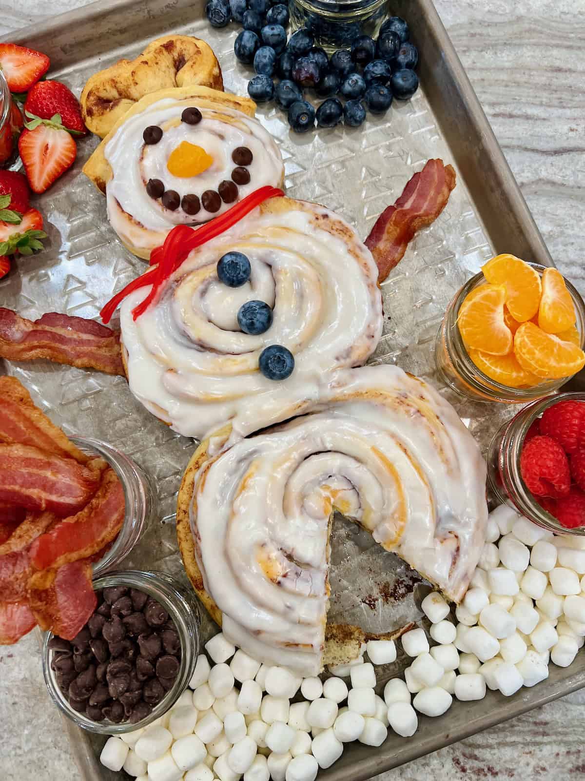 A snowman made of three cinnamon rolls on a baking sheet surrounded by mason jars filled with bacon, strawberries and mandarin slices.