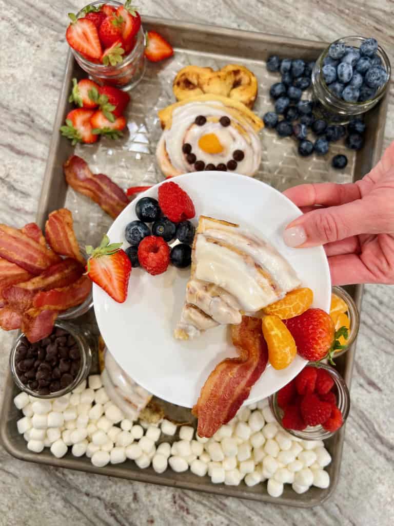 A white plate with a slice of cinnamon roll, mixed berries, bacon and mandarin orange slices.