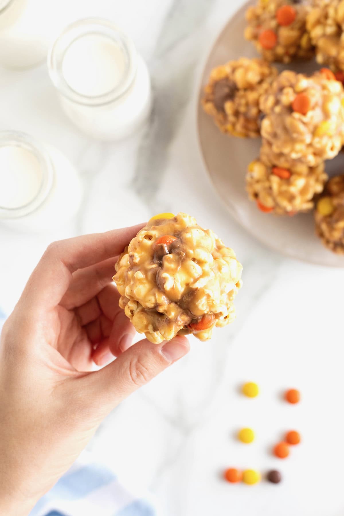 A hand holding a peanut butter popcorn ball in from of a large white serving plate of popcorn balls.