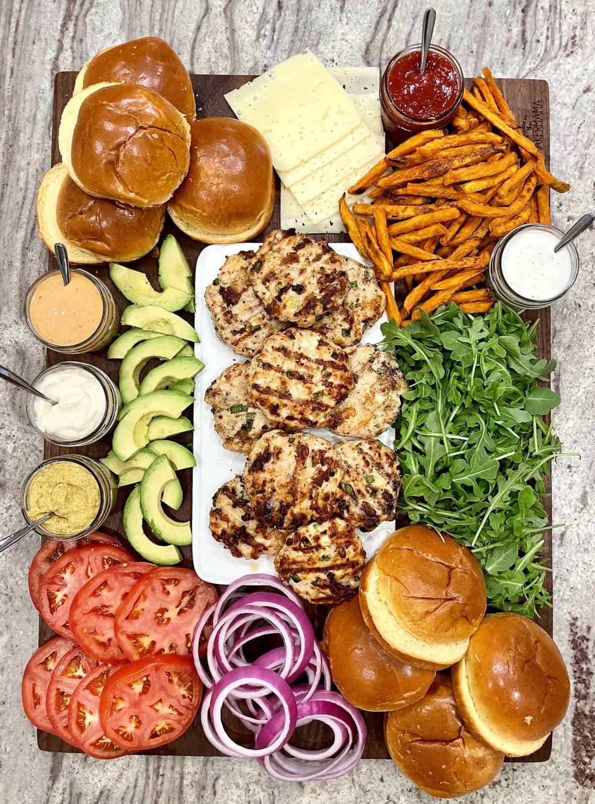 Father's Day grilled turkey burgers, homemade sweet potato fries sliced avocado, sliced tomato, red onion slices and jars of sauces served on a dark wood food board resting on a gray marble counter.