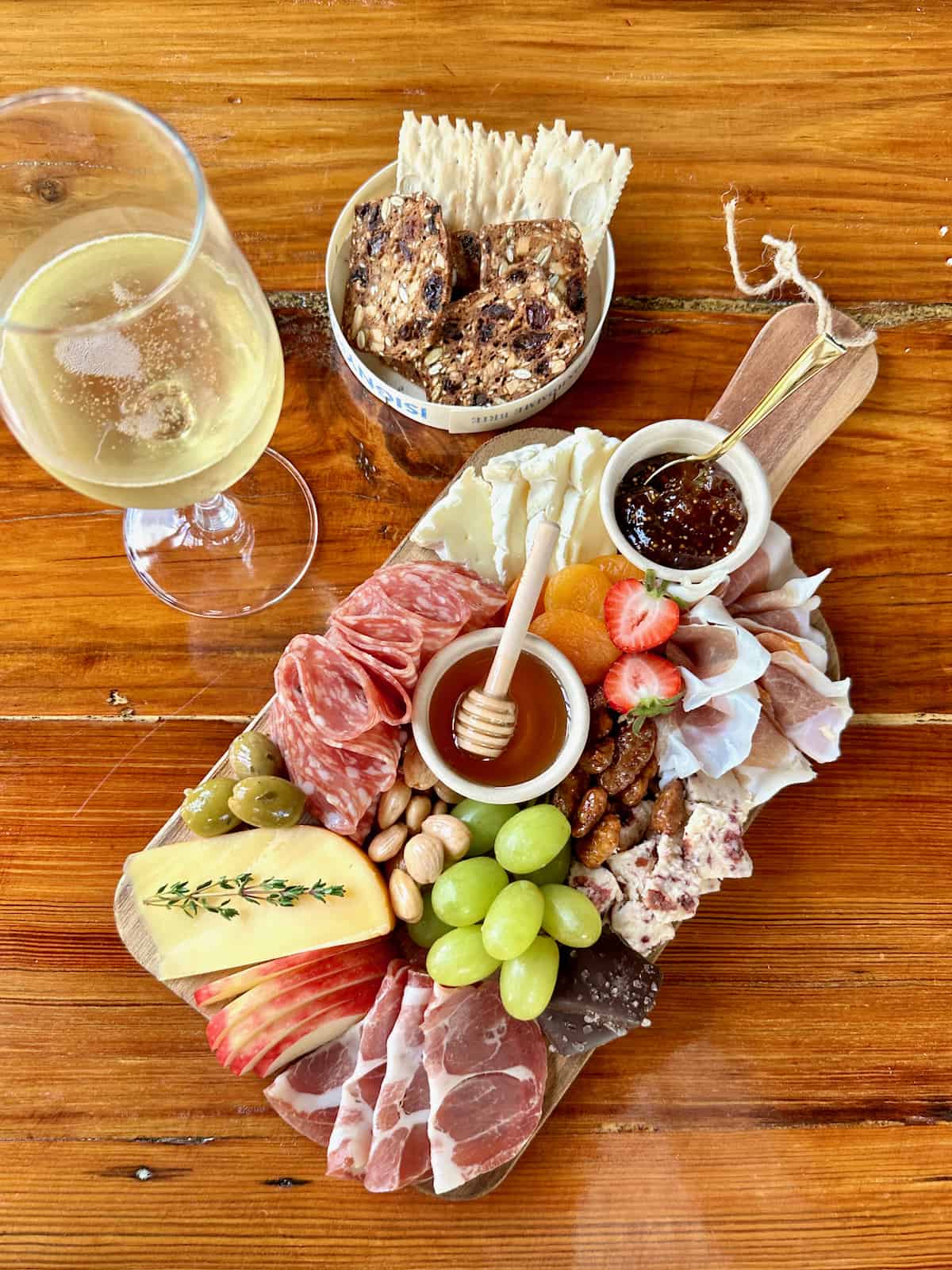 A natural wood serving board with fresh green grapes, apples, honey and a honey diaper next to a glass of white wine.