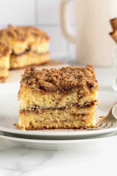 A square slice of coffee cake on a stack of white ceramic plates with a metal fork.