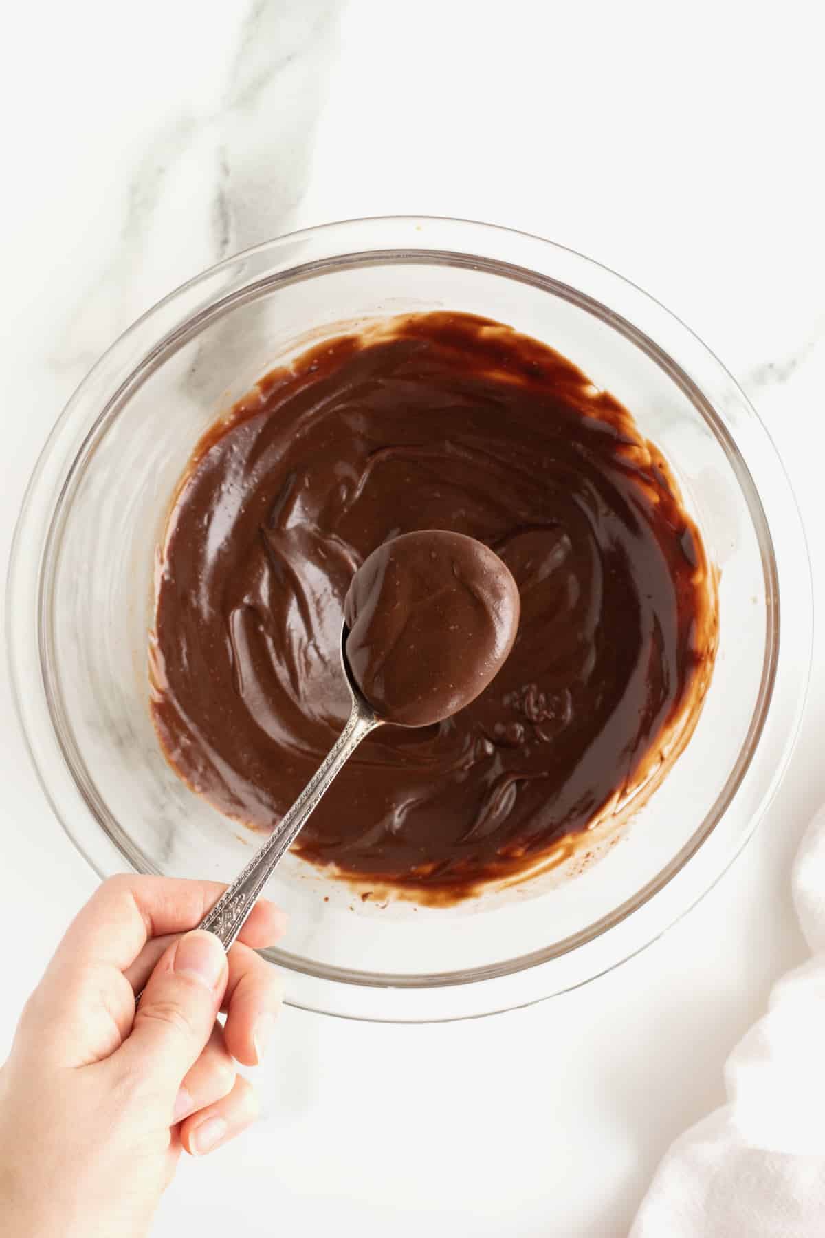 Chocolate ganache in a clear glass bowl with a hand holding a spoonful of chocolate over it.