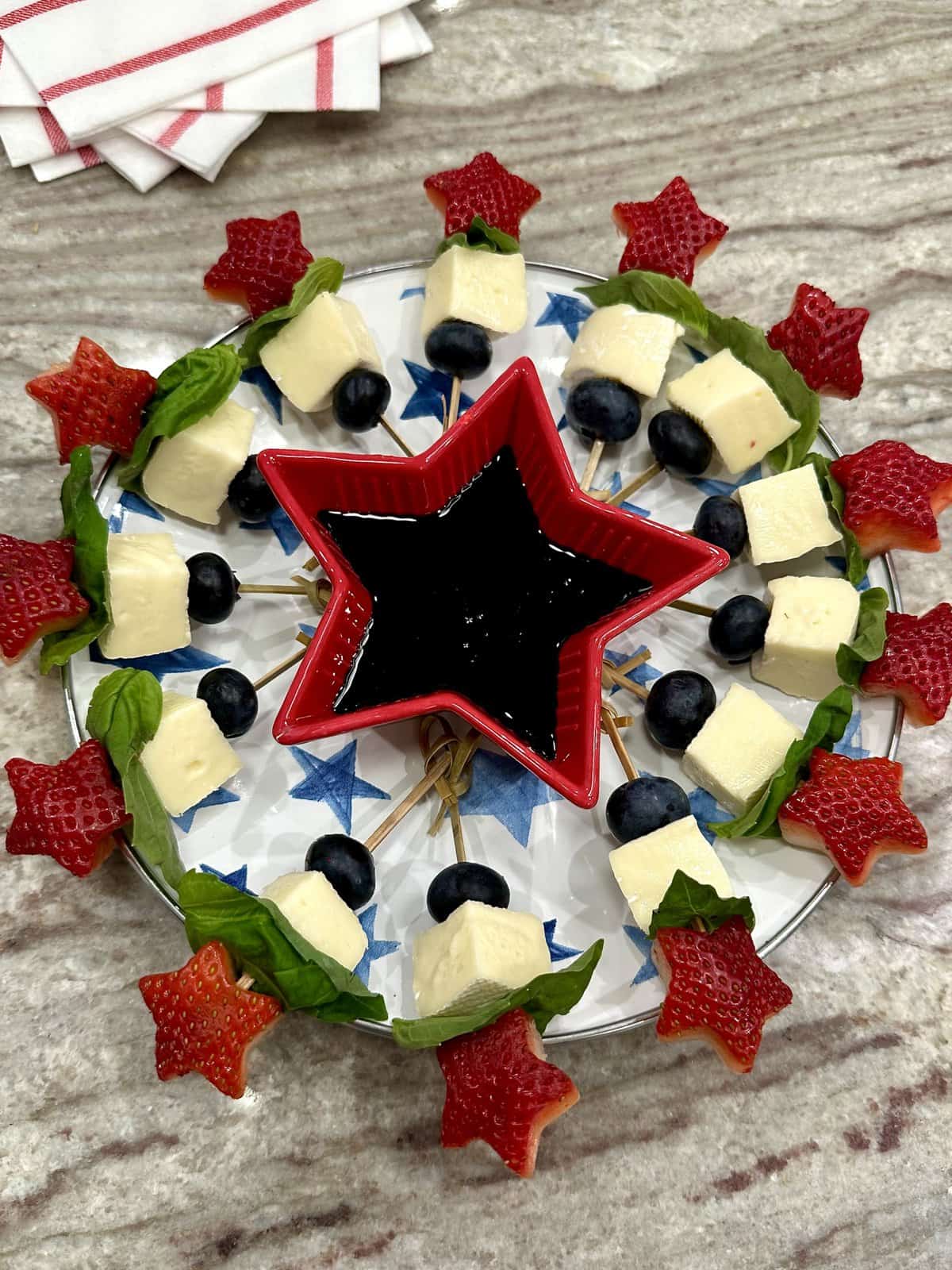 Star-shaped strawberry halves on fruits skewer with chunks of brie and blueberries surrounding a red star bowl filled with balsamic glaze.