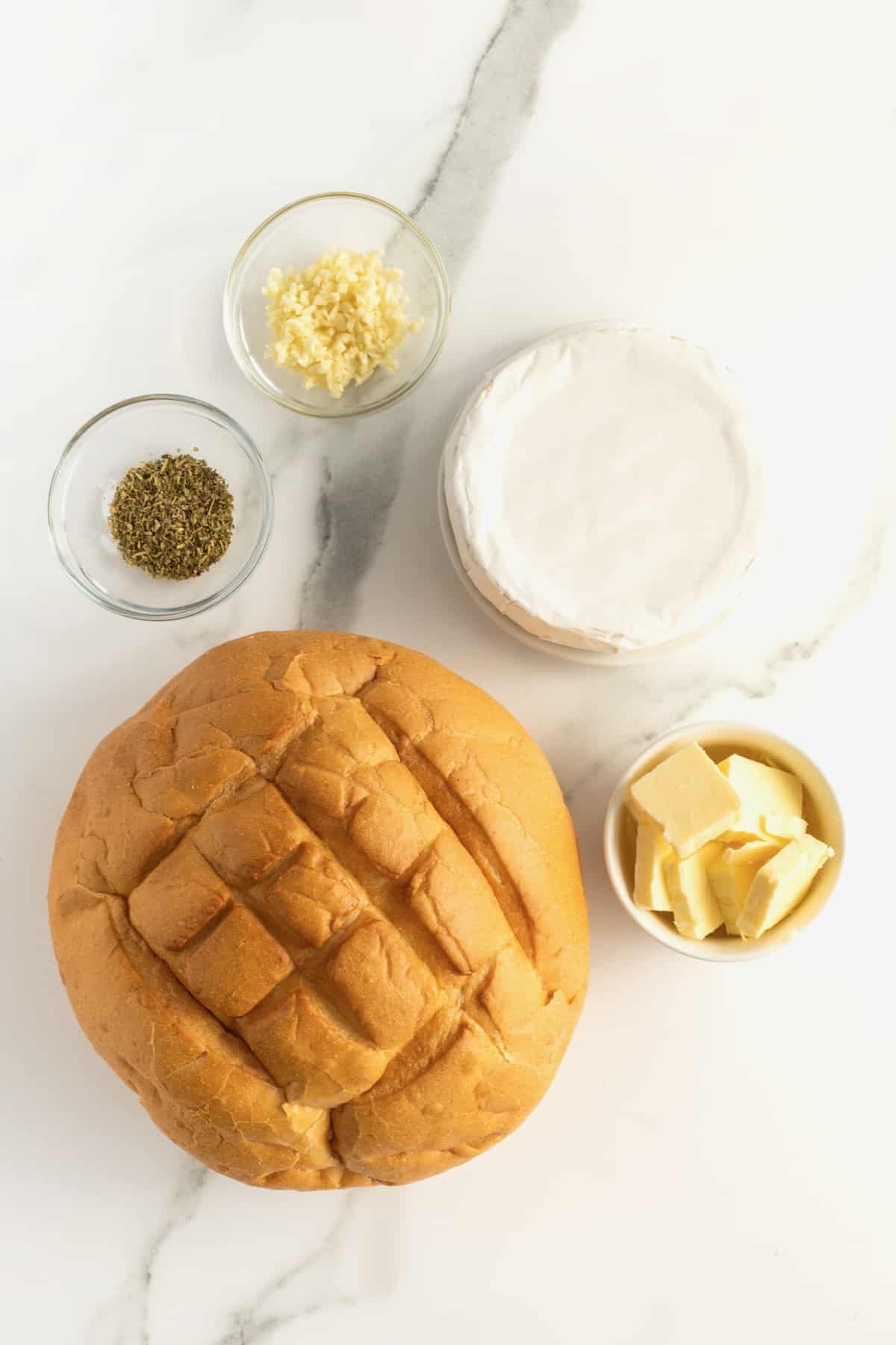 A bread bowl on a marble countertop, next to a wheel of brie and glass bowls filled with garlic, butter, and Italian seasoning.