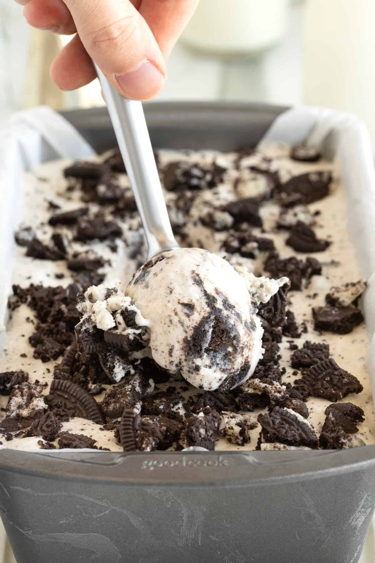 A metal spoon scooping Cookies n' Cream ice cream out of a loaf pan lined with parchment paper.