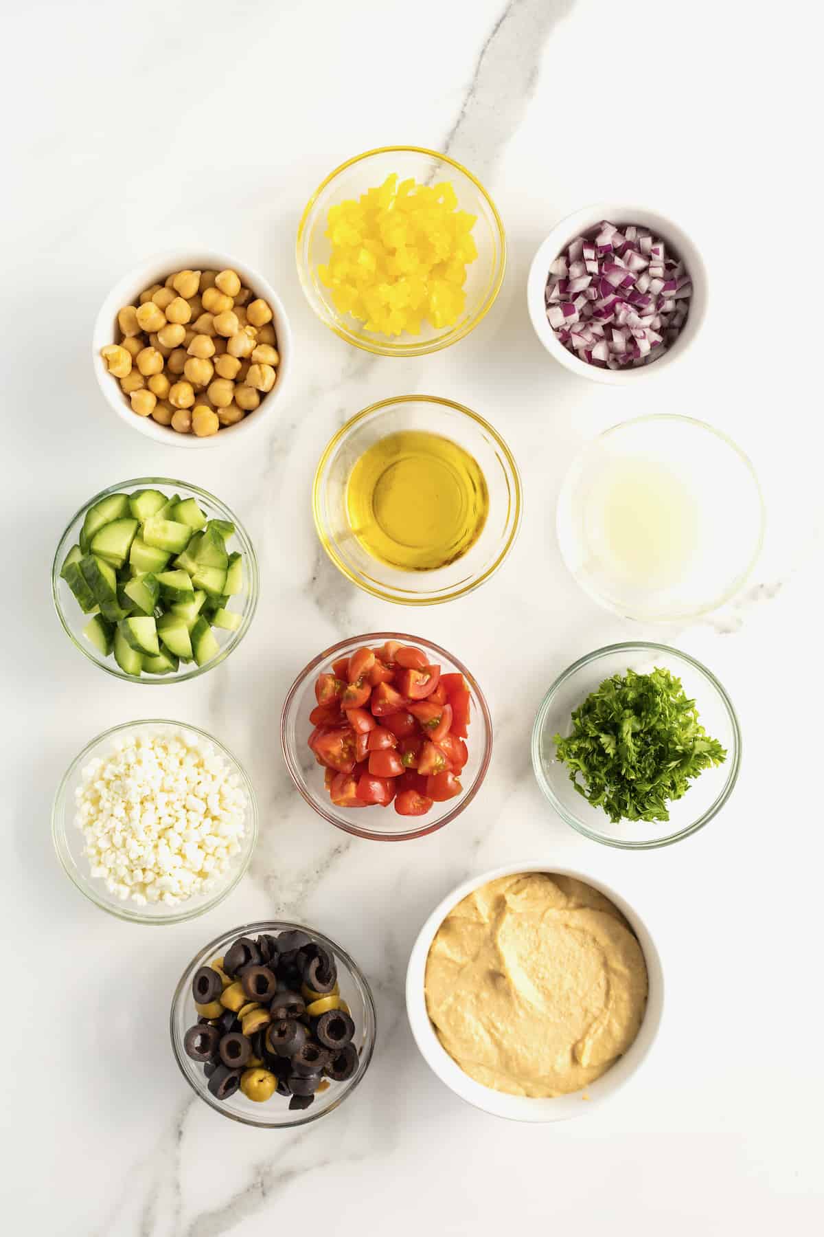 Tomatoes, olives, cucumber, chickpeas, red onion and pepperoncini peppers in glass and ceramic dishes and hummus in a white ceramic dish.