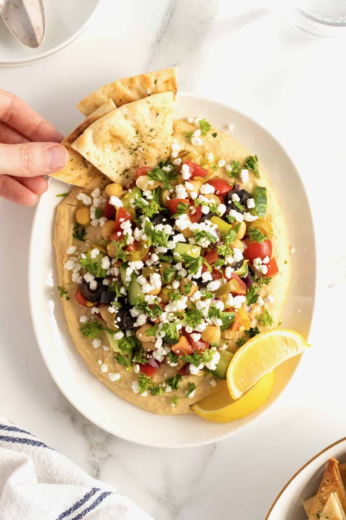 Hand grabbing pita triangle from loaded hummus topped with tomatoes, olives, cucumber, chickpeas, red onion and pepperoncini peppers in a white ceramic serving dish.