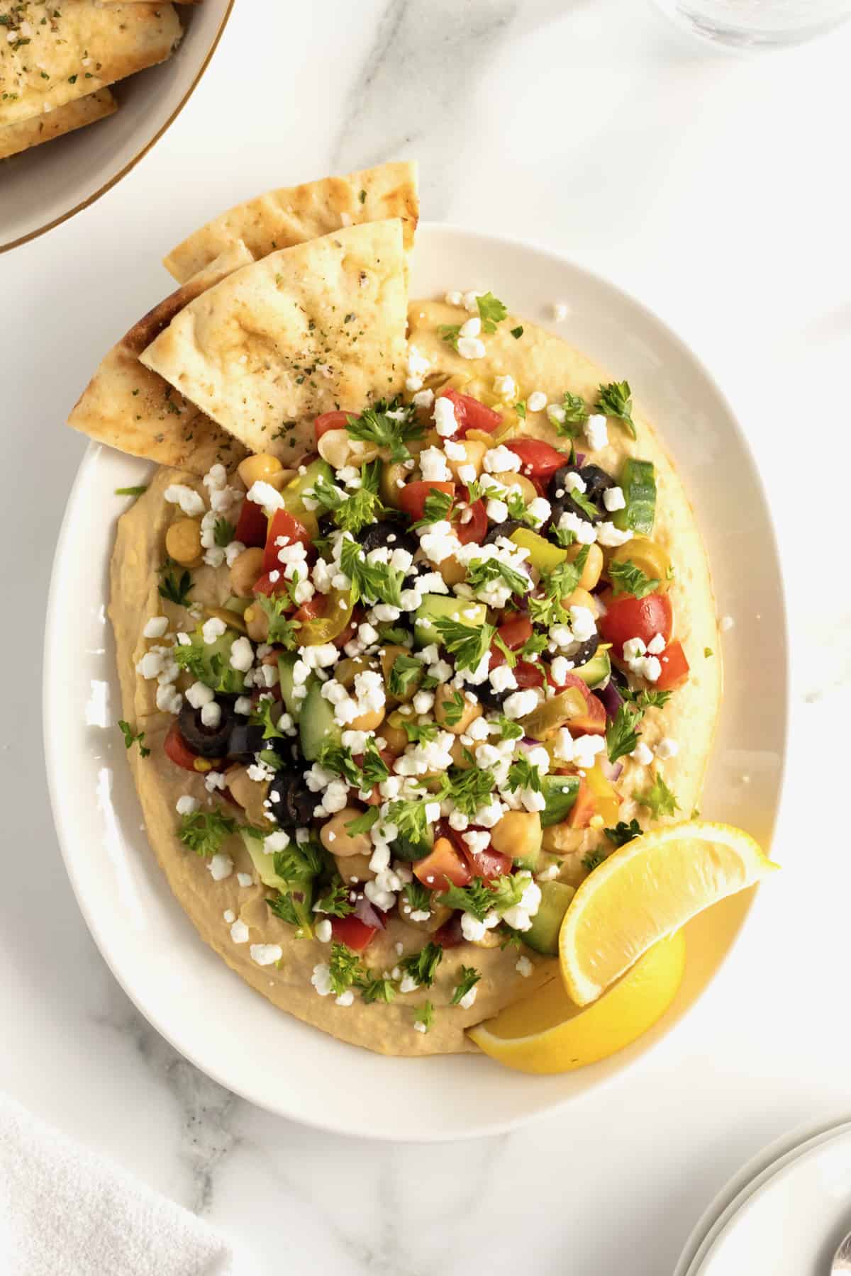 Hummus topped with tomatoes, olives, cucumber, chickpeas, red onion and pepperoncini peppers in a white ceramic serving dish with pita triangles.