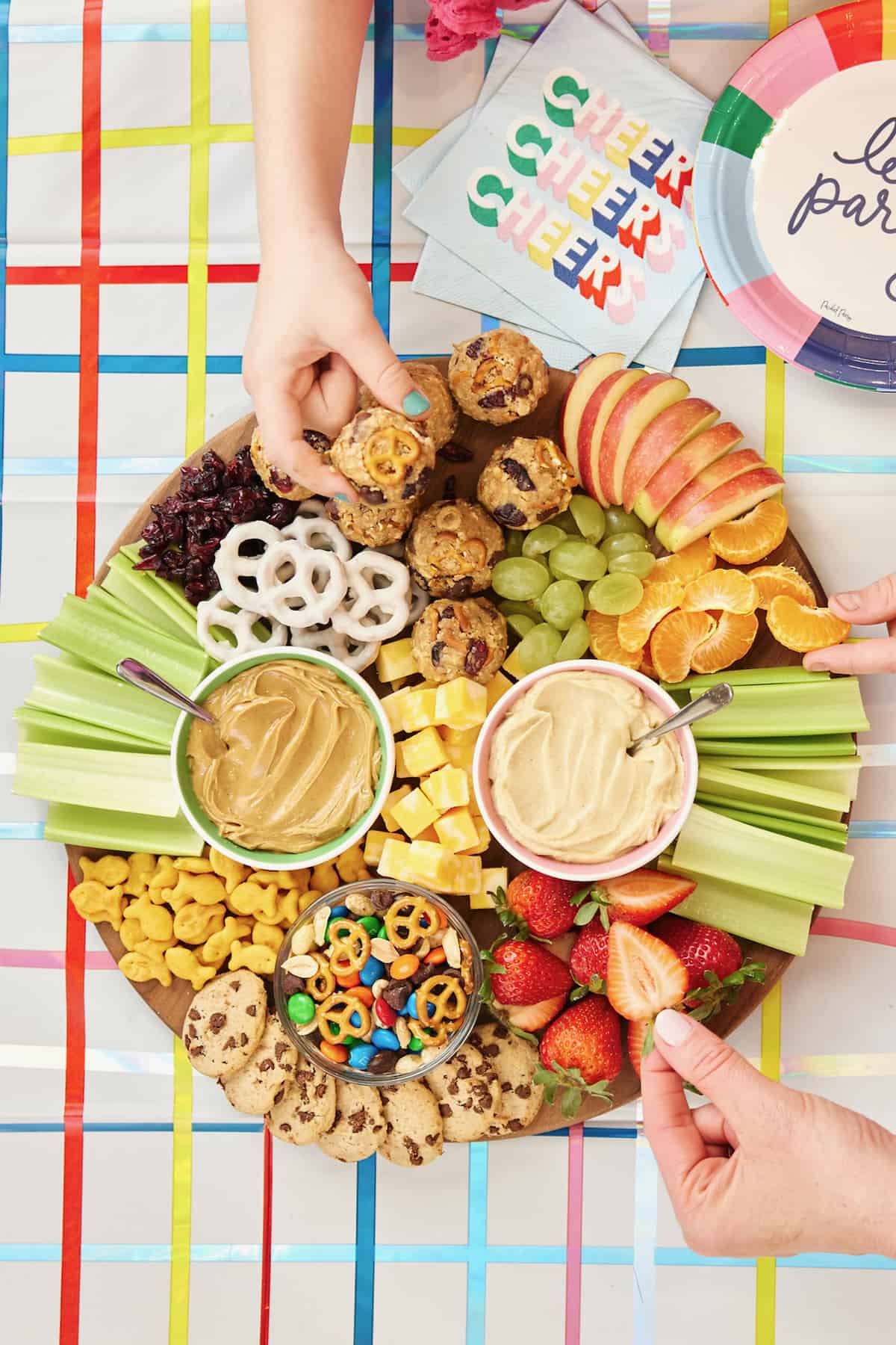 A round wood board with Hummus, Peanut butter, Snack Mix (made with peanuts, mini pretzels, chocolate chips and M&M’s), Cheese cubes, Celery pieces, Apple slices, Mandarin orange segments, Green grape halves, Strawberry halves, Goldfish crackers, Dried cranberries, Yogurt covered pretzels and Mini chocolate chip cookies.