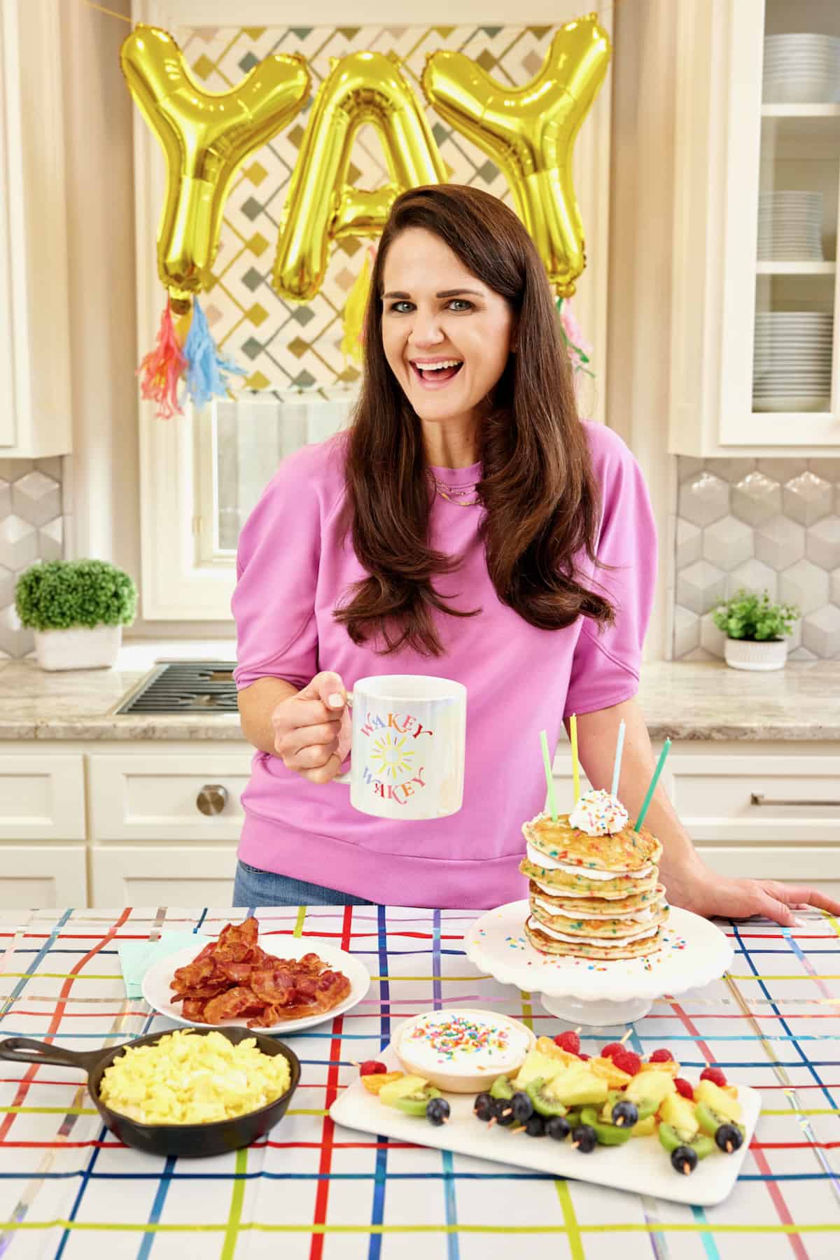 A breakfast spread featuring soft scrambled eggs in black cast iron skillet, bacon on a round white plate, a stack of confetti pancakes on a white cake plate, and a pile of fruit skewers made with raspberries, mandarin oranges, pineapple pieces and a blueberry on a rectangular white serving tray.