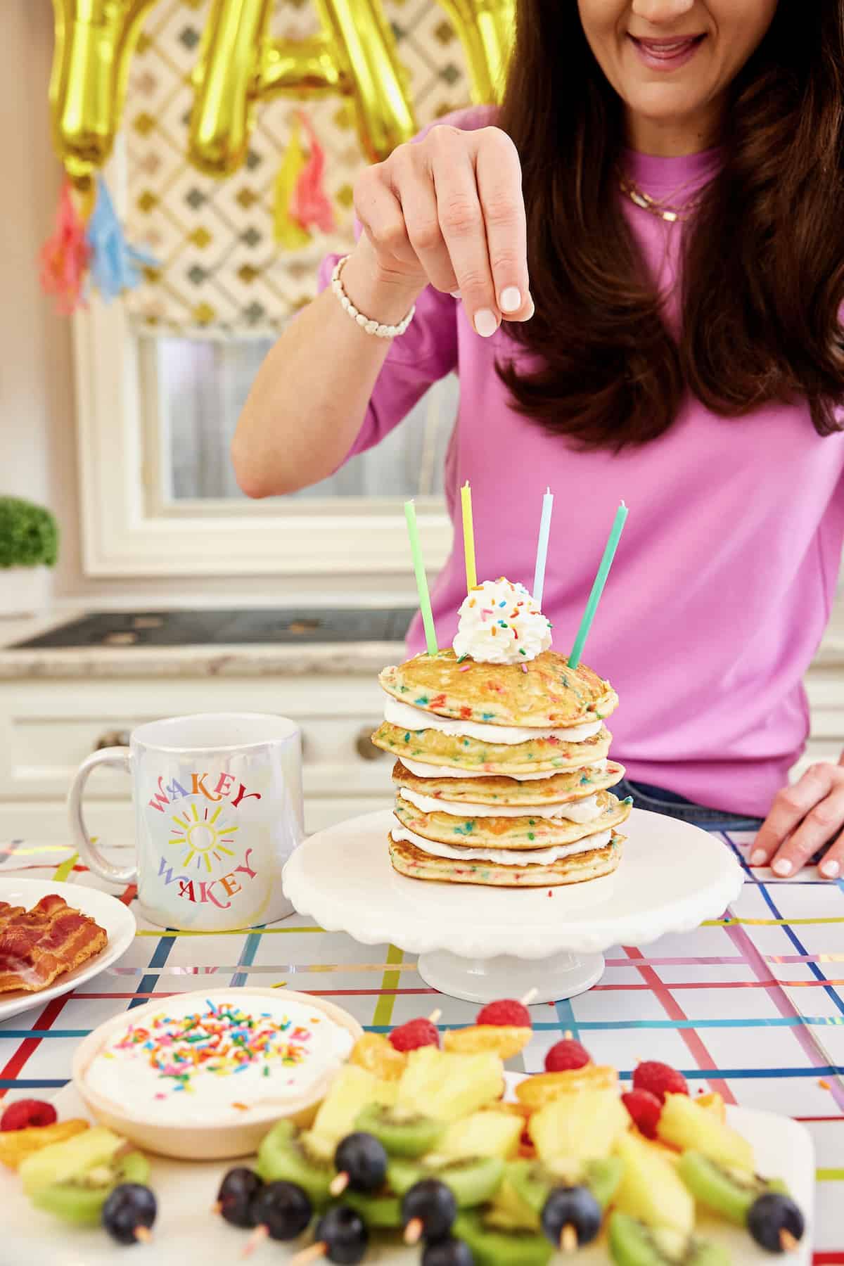 The BakerMama sprinkling rainbow sprinkles onto stack of five pancakes with sprinkles baked in and whipped topping between each layer and four candles on top.