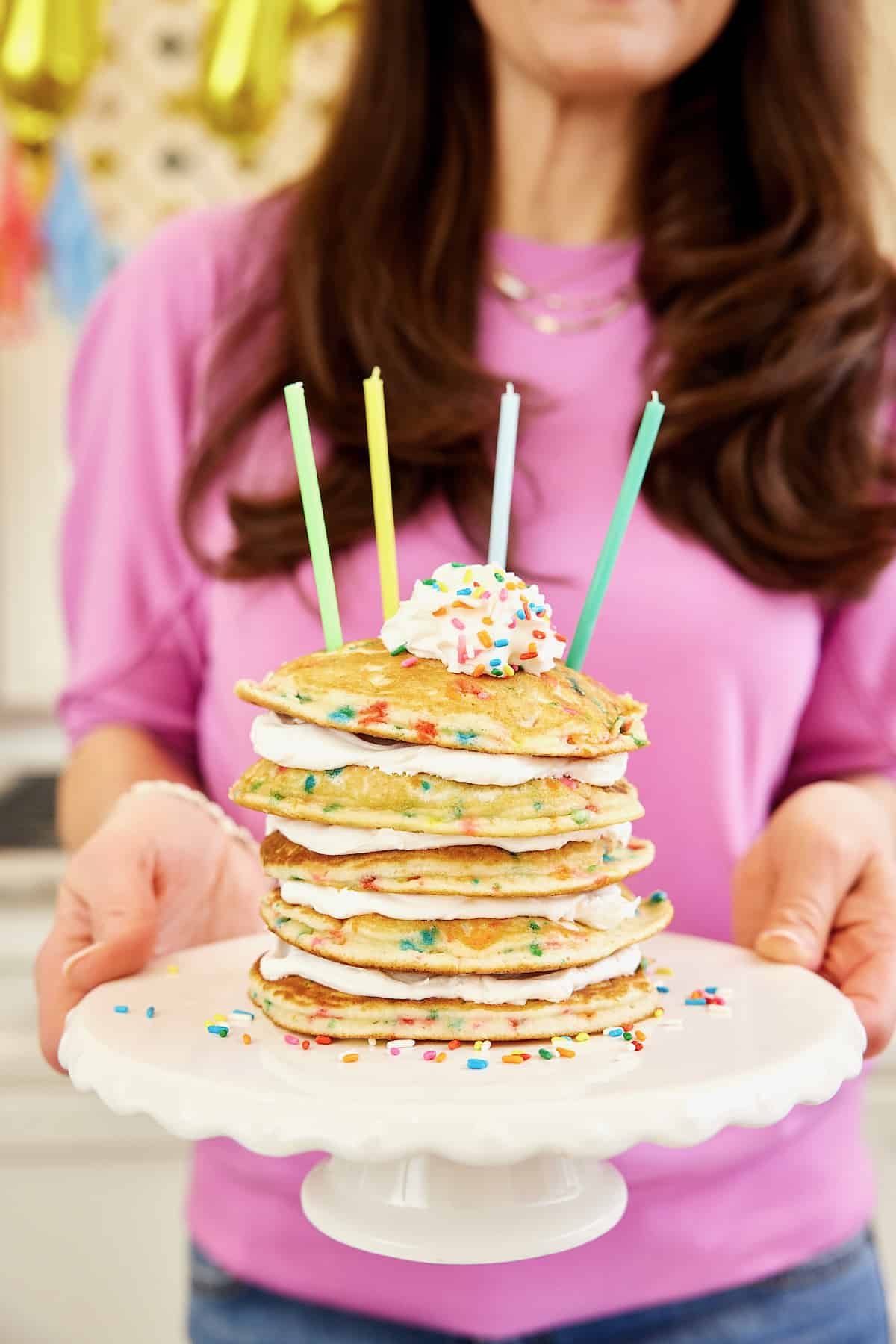 A stack of five confetti pancakes with layers of whipped topping between on a white cake plate with four candles on top.