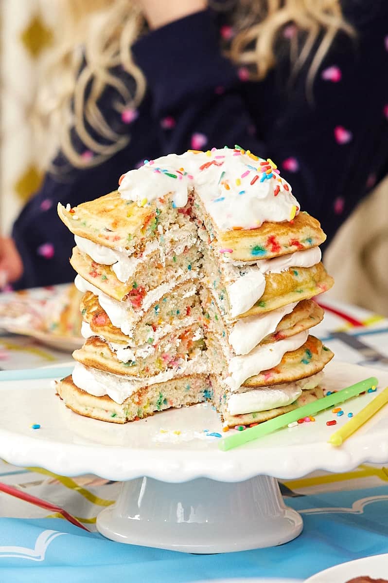Whipped topping between five layers of confetti pancakes on a white cake plate.
