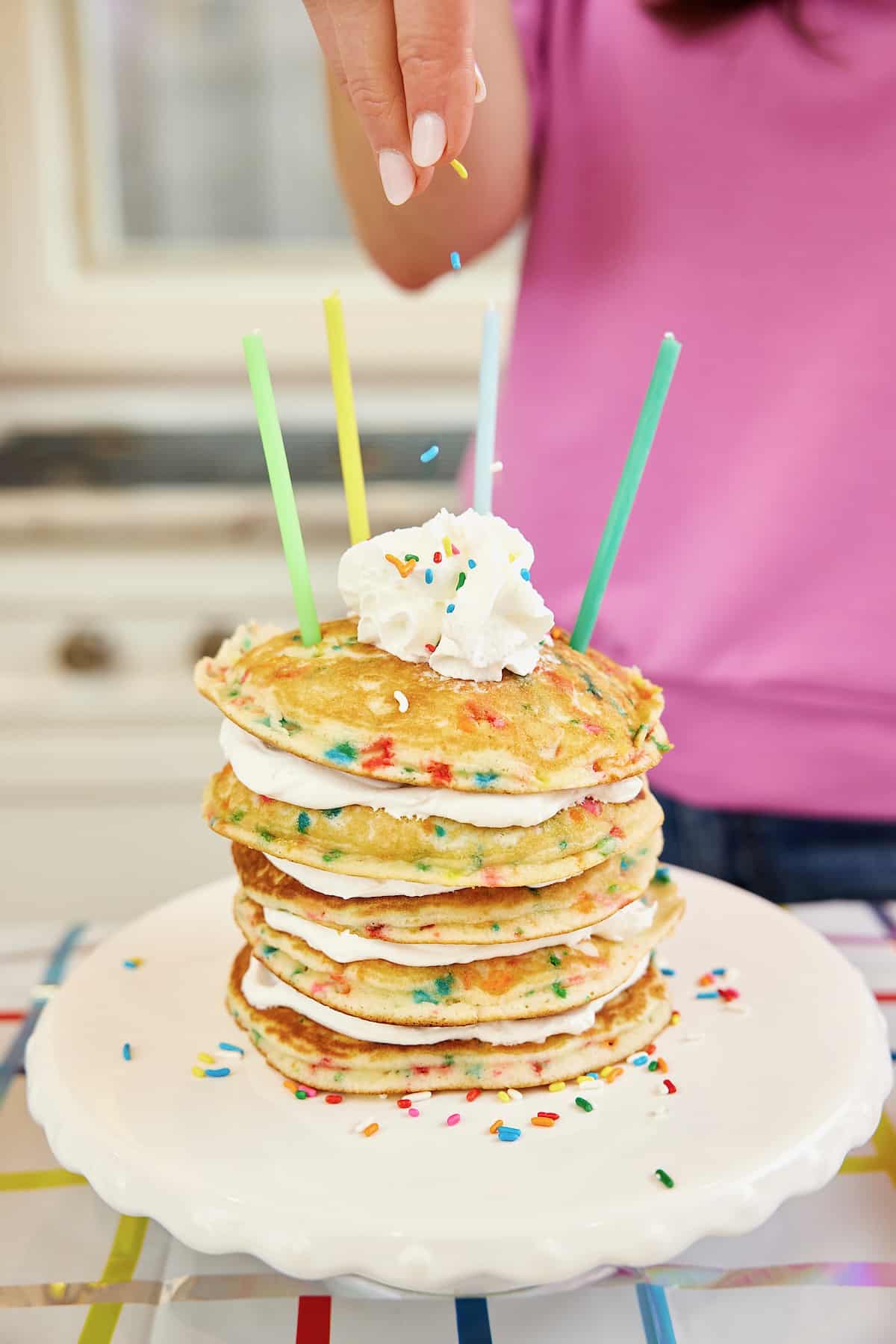 Sprinkles being dropped onto a stack of five pancakes with sprinkles with whipped topping between each layer and four candles on top.