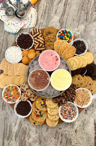 A large wooden board with chocolate, strawberry and vanilla ice creams, cookies and treats like mini marshmallows, M&Ms, crushed Oreos, rainbow sprinkles, Fruity Pebbles and crushed peanuts.