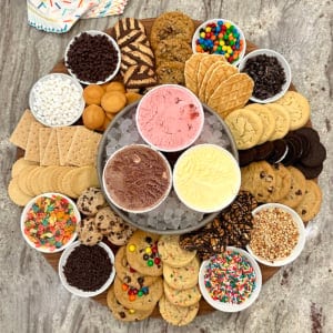 A large wooden board with chocolate, strawberry and vanilla ice creams, cookies and treats like mini marshmallows, M&Ms, crushed Oreos, rainbow sprinkles, Fruity Pebbles and crushed peanuts.