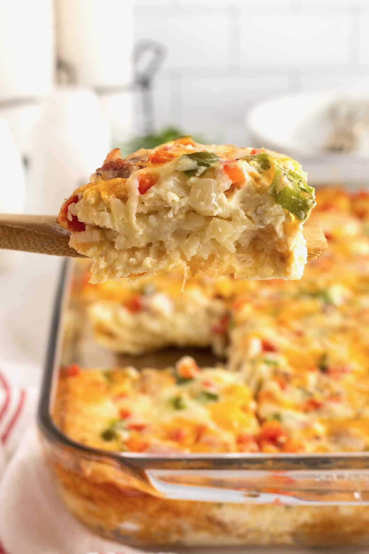 A slice of breakfast casserole on a wooden spatula with a glass baking dish of Hash Brown Breakfast Casserole in the background.