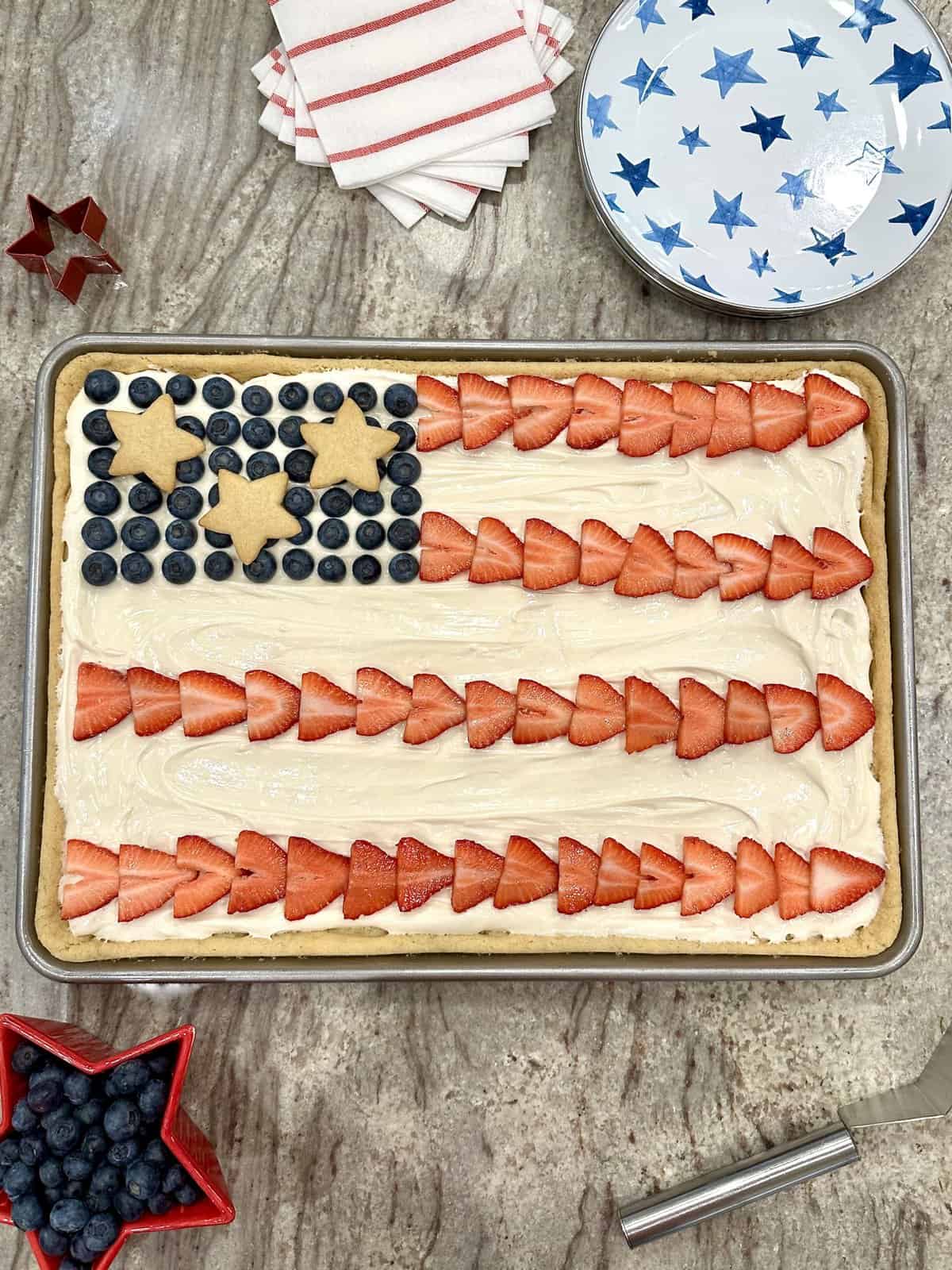 A rimmed cookie sheet with a large sugar cookie frosted and decorated like an American Flag using sliced strawberries, fresh blueberries and star cookies.