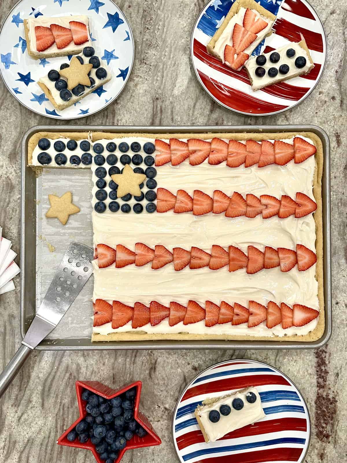 A rimmed cookie sheet with a large sugar cookie frosted and decorated like an American Flag using sliced strawberries, fresh blueberries and star cookies.