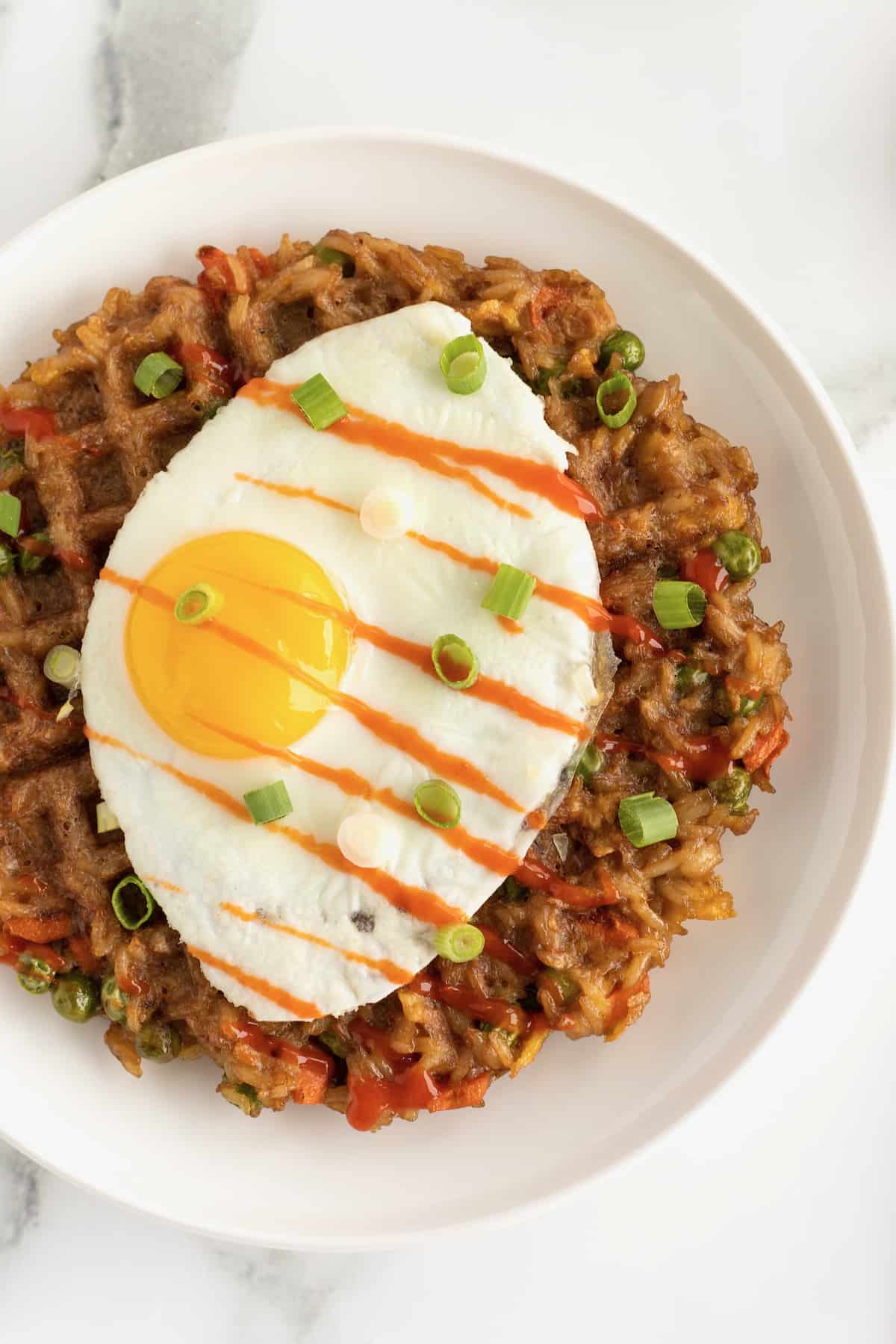 A sunny side up egg on top of a fried rice waffle on a white ceramic plate sprinkled with chopped green onion and drizzle with sriracha sauce.