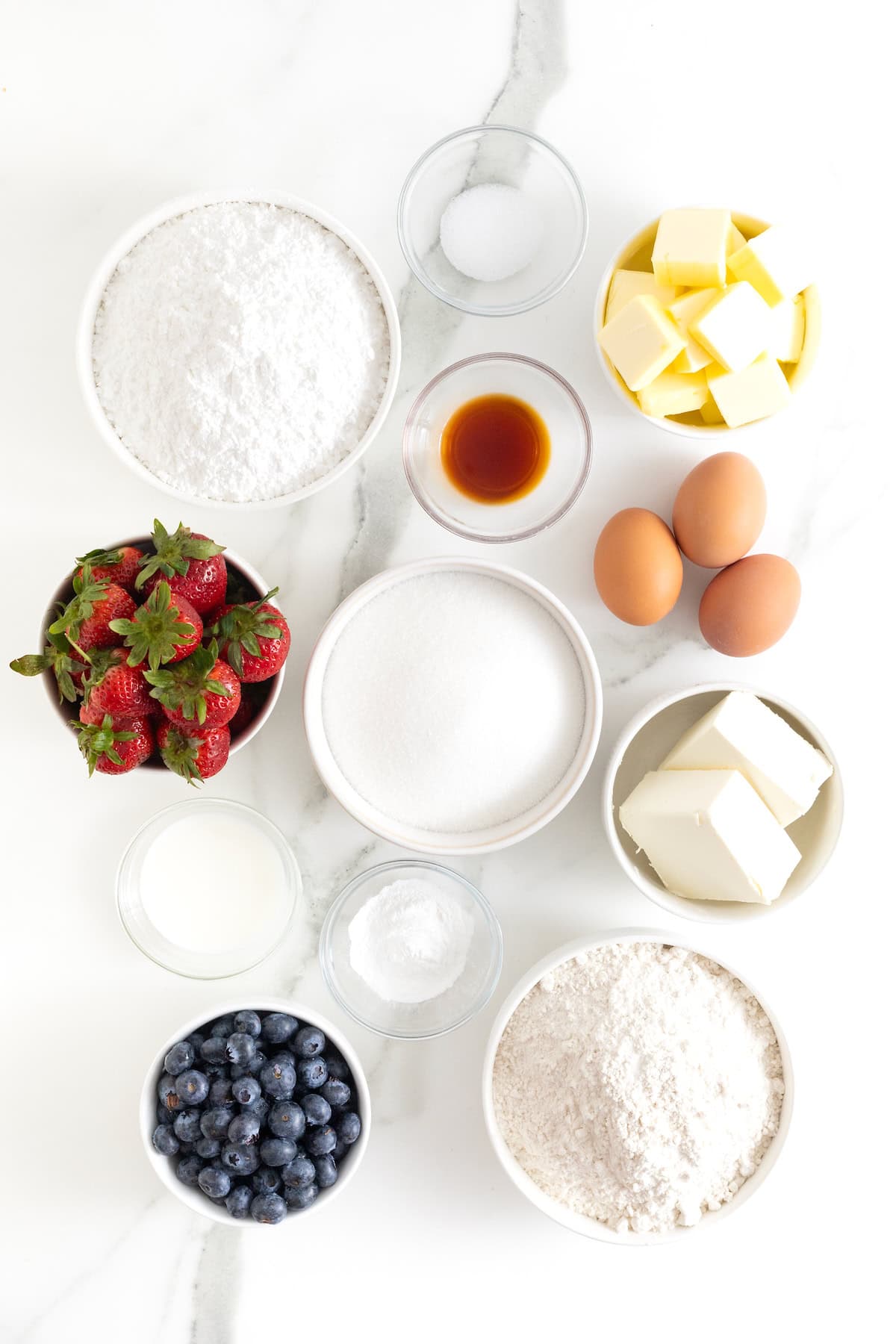 Ingredients to make American Flag Fruit Pizza in small glass dishes on a white marble counter.