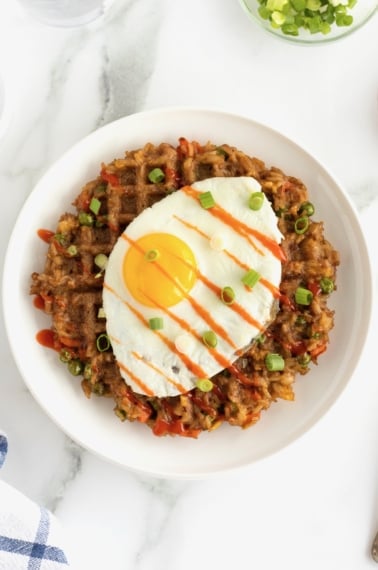 A sunny side up egg on to p of a fried rice waffle on a white ceramic plate.