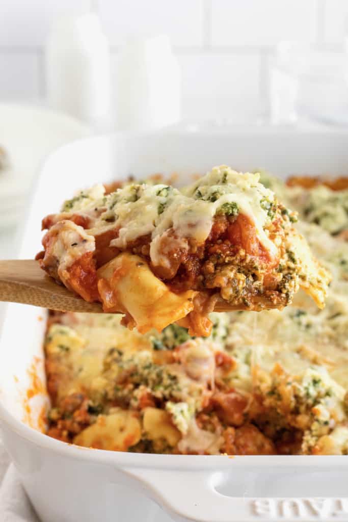 A wooden spoon holding pasta covered in tomato sauce, melted mozzarella cheese and fresh herbs over a white casserole dish of lasagna.
