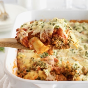 A wooden spoon holding pasta covered in tomato sauce, melted mozzarella cheese and fresh herbs over a white casserole dish of lasagna.