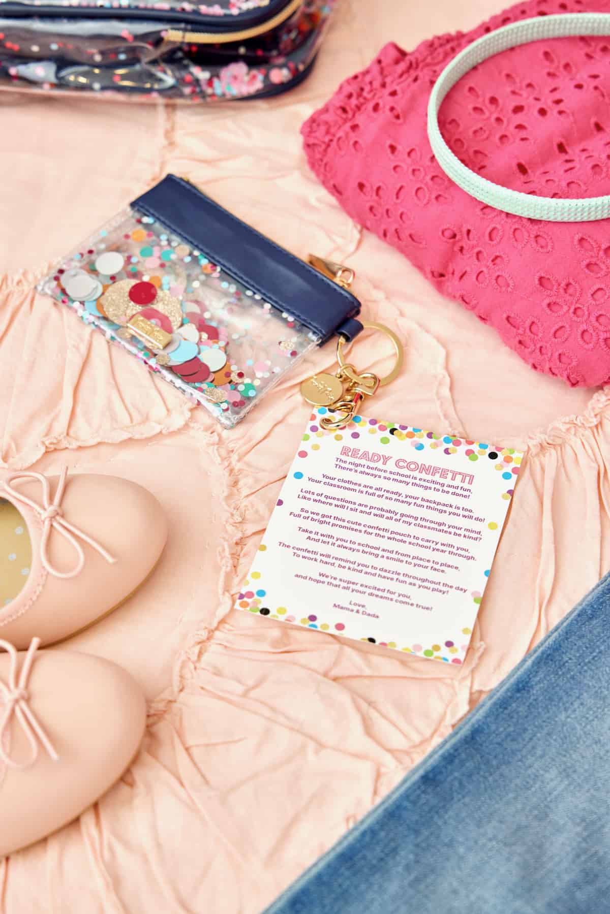 A small key chain pouch with glitter, a pair of child's pink ballet flats, and a bright pink eyelet lace child's shirt on a pale pink ruffled bedspread.