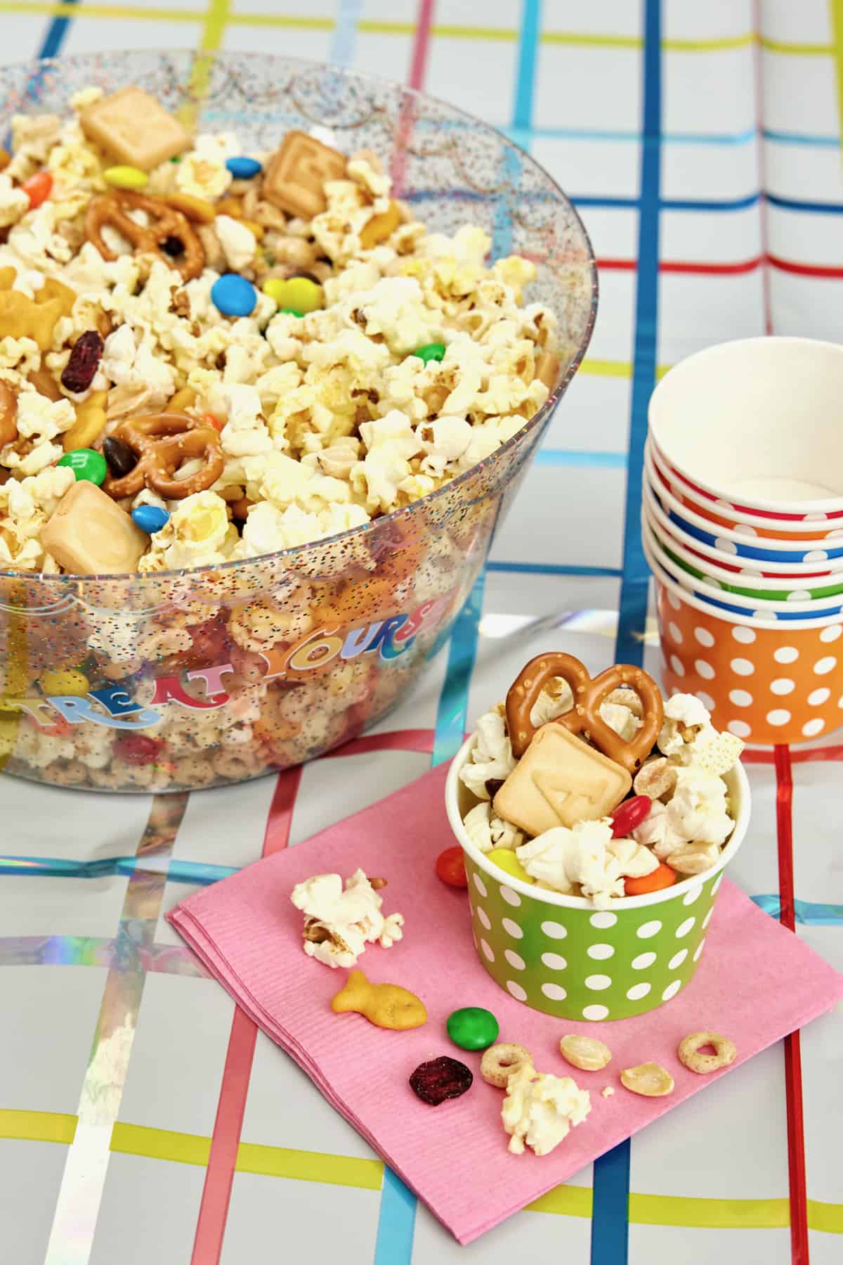 A stack of paper cups next to a large glass bowl reading TREAT YOURSELF filled with a popcorn snack mix with pretzels, M&Ms, dried cranberries, roasted peanuts and alphabet cookies.