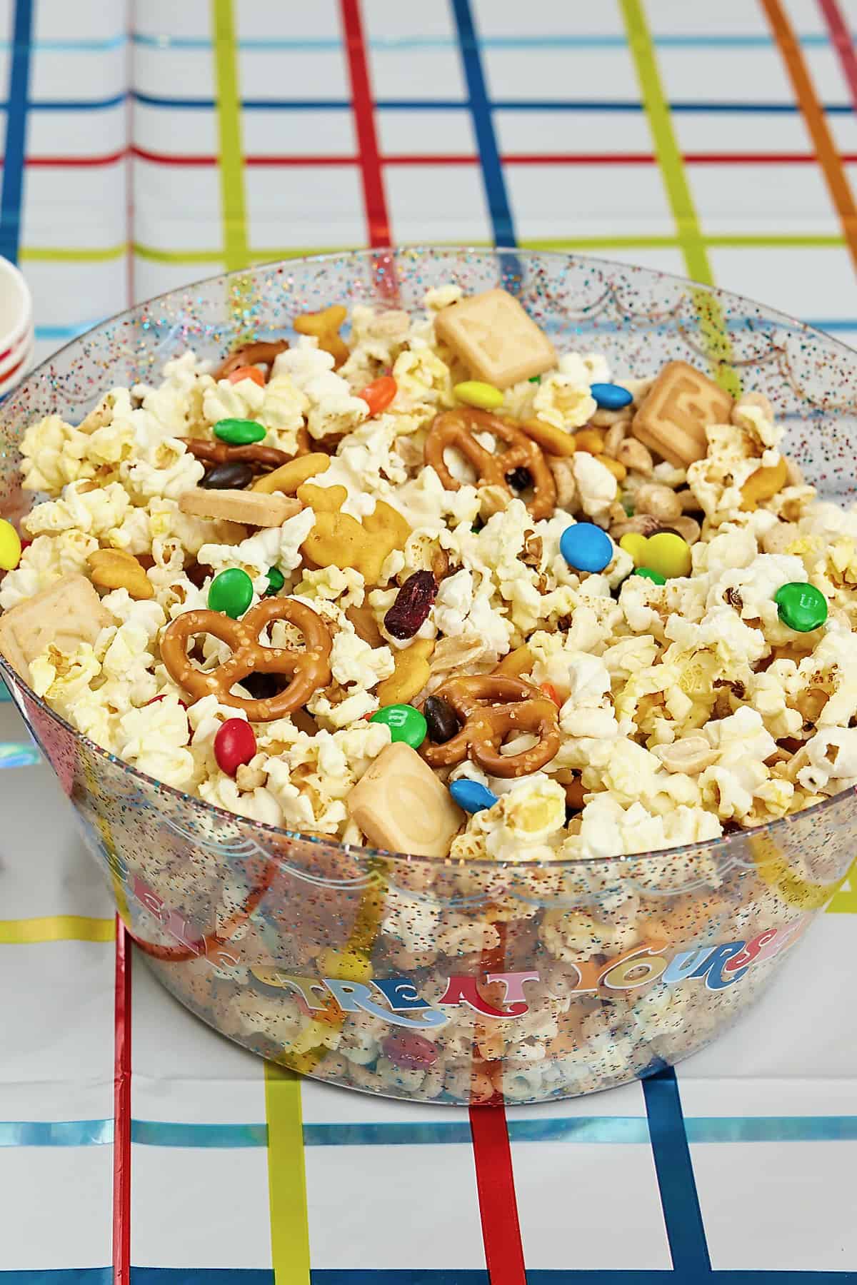 A large glass bowl reading TREAT YOURSELF filled with a popcorn snack mix with pretzels, M&Ms, dried cranberries, roasted peanuts and alphabet cookies.