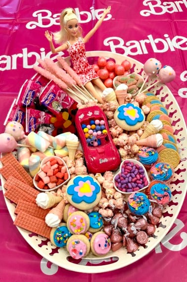 A Barbie dessert tray with pink cookies, popcorn, cake pops and a Barbie doll on a Barbie themed table cloth.