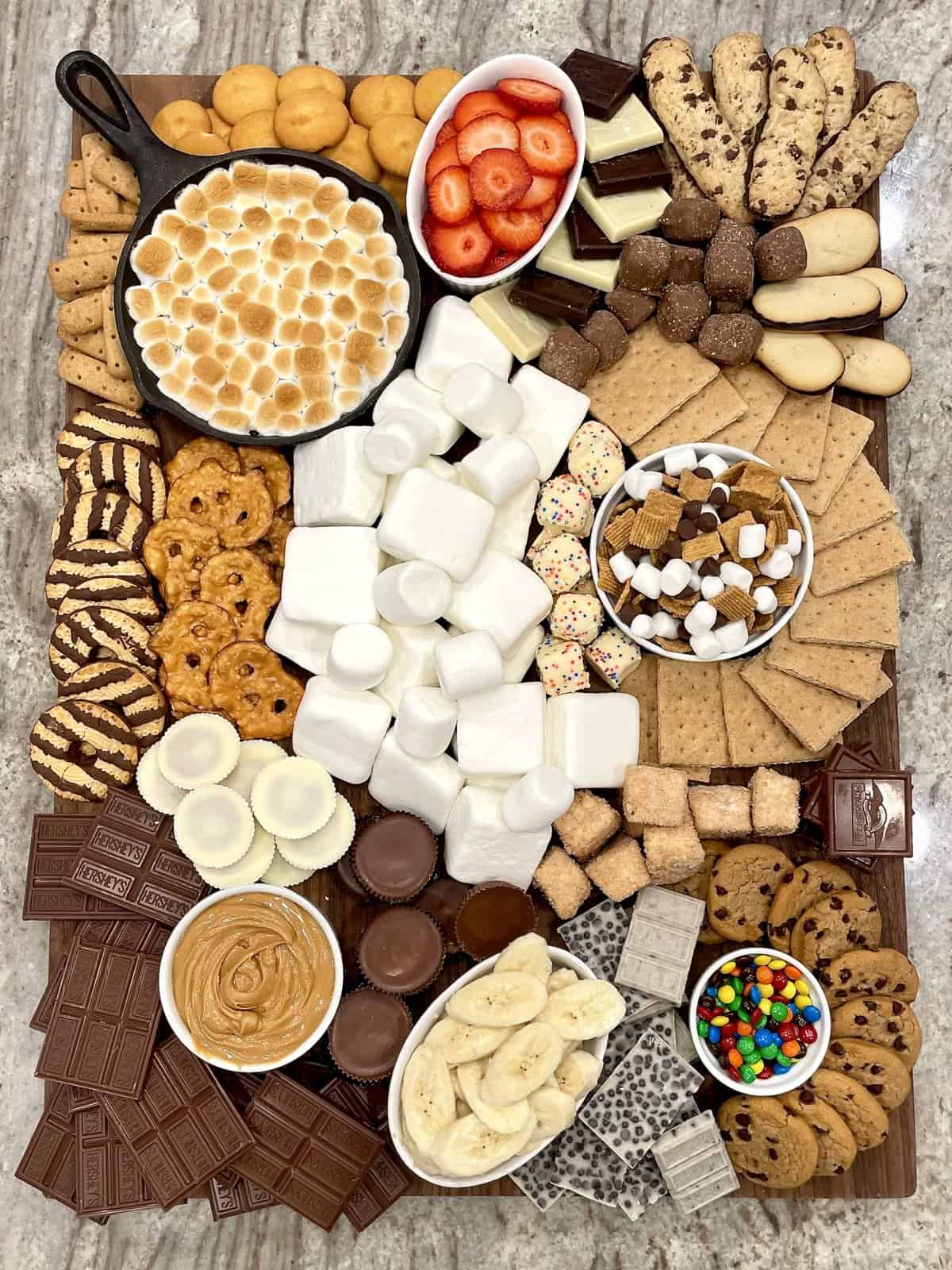 A large wooden board with graham crackers, marshmallows, Fudge Stripe cookies, S'mores skillet dip, sliced strawberries and bananas, pretzel chips, and chocolate chip cookies.