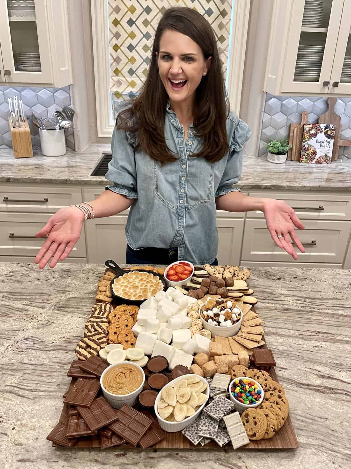 A large wooden board with graham crackers, marshmallows, Fudge Stripe cookies, S'mores skillet dip, sliced strawberries and bananas, pretzel chips, and chocolate chip cookies.