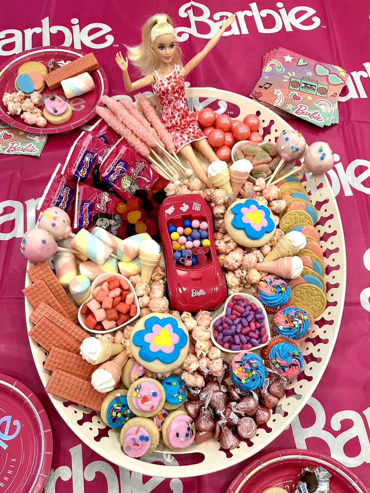 A Barbie dessert tray with pink cookies, popcorn, cake pops and a Barbie doll on a Barbie themed table cloth.